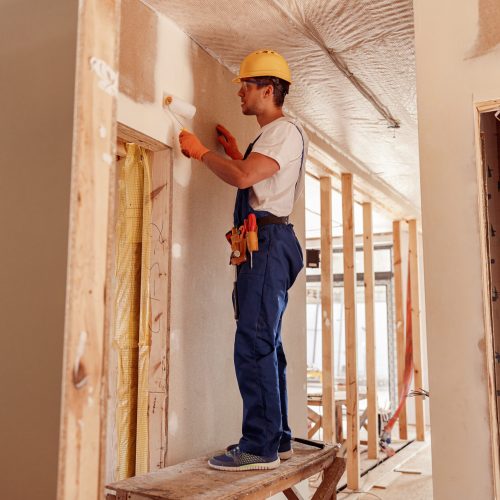 Handsome young man in safety helmet and work overalls painting wall with paint roller